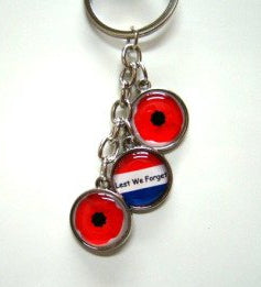 Metalen sleutelhanger met drie ronde bedeltjes, twee met rode klaprozen en één met de tekst Lest We Forget op een achtergrond met de Nederlandse vlag.